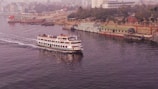 A passenger ferry is moving along a calm river, with a number of people on board enjoying the view. The background features a cityscape with several buildings and a densely wooded area. On the opposite bank, a few moored ships and industrial structures are visible.