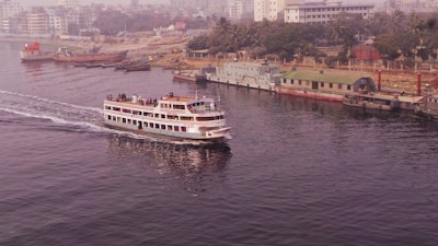 A passenger ferry is moving along a calm river, with a number of people on board enjoying the view. The background features a cityscape with several buildings and a densely wooded area. On the opposite bank, a few moored ships and industrial structures are visible.