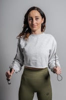 A woman is standing while holding a jump rope. She is wearing a gray cropped sweatshirt and olive green leggings. Her hair is loose and wavy, and she has a slight smile on her face.