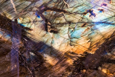 A close-up view of a polished mineral surface displaying an iridescent play of colors. The stone's internal lines and fractures enhance the visual complexity, with hues ranging from blues and greens to browns and golds.