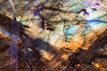 A close-up view of a polished mineral surface displaying an iridescent play of colors. The stone's internal lines and fractures enhance the visual complexity, with hues ranging from blues and greens to browns and golds.