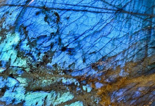 An abstract image showcasing a close-up of a labradorite mineral, featuring an intricate pattern of blue and gold iridescent colors. The surface is cracked with lines that create a natural network, reflecting light and displaying varying shades of blue and green with hints of yellow. The surface texture appears smooth yet irregular, with some darker spots adding depth.