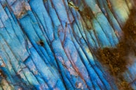 A close-up view of a mineral or gemstone surface displaying vibrant blue hues with streaks of dark lines and patches of brown and green. The surface texture is glossy and has an iridescent quality, with subtle variations in color and pattern throughout.