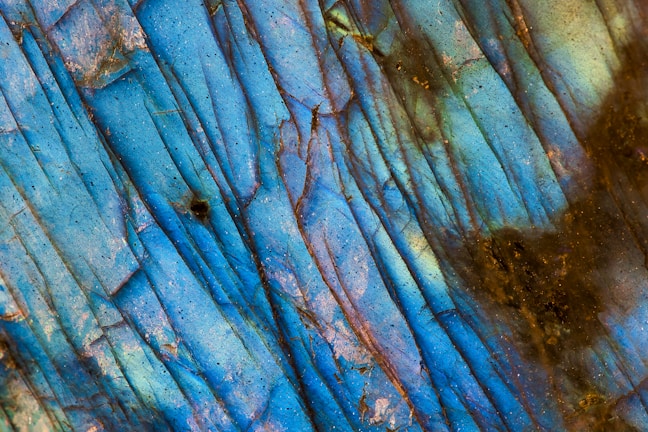 A close-up view of a mineral or gemstone surface displaying vibrant blue hues with streaks of dark lines and patches of brown and green. The surface texture is glossy and has an iridescent quality, with subtle variations in color and pattern throughout.