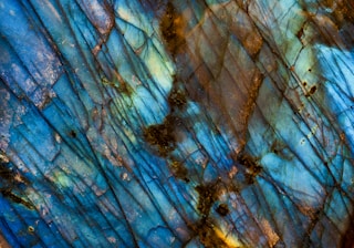 A close-up view of a labradorite mineral with vibrant iridescent colors displayed on its surface. The crystal structure reveals a range of blue and green hues with hints of brown and orange, creating an intricate pattern of overlapping lines and textures.