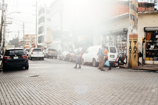 A busy urban street scene with several people walking across the road. Cars are parked and moving along the street, which is lined with buildings showcasing various storefronts. A yellow pillar with 'TAXI' signage stands prominently on the sidewalk.