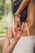 A woman in a white tank top performs a yoga stretch by interlocking her fingers behind her back. Her right arm is bent upward while the left arm reaches downward. She wears a bracelet made of colorful beads. The background shows a blurred view of greenery, indicating an outdoor setting.
