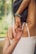 A woman in a white tank top performs a yoga stretch by interlocking her fingers behind her back. Her right arm is bent upward while the left arm reaches downward. She wears a bracelet made of colorful beads. The background shows a blurred view of greenery, indicating an outdoor setting.
