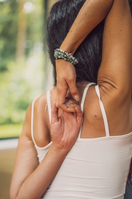 A woman in a white tank top performs a yoga stretch by interlocking her fingers behind her back. Her right arm is bent upward while the left arm reaches downward. She wears a bracelet made of colorful beads. The background shows a blurred view of greenery, indicating an outdoor setting.