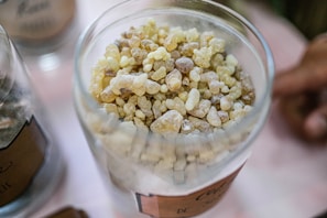 A close-up view of a glass container filled with small, irregularly shaped, pebble-like granules in shades of yellow, brown, and off-white. The container is situated on a light-colored surface with another similar container partially visible on the side.