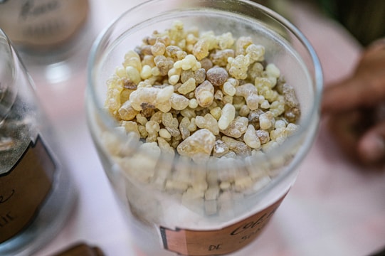 A close-up view of a glass container filled with small, irregularly shaped, pebble-like granules in shades of yellow, brown, and off-white. The container is situated on a light-colored surface with another similar container partially visible on the side.