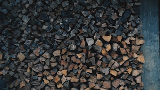 A stacked pile of chopped firewood in various shades of brown, neatly arranged against a wooden backdrop. The firewood pieces are of different shapes and sizes, creating a textured pattern.