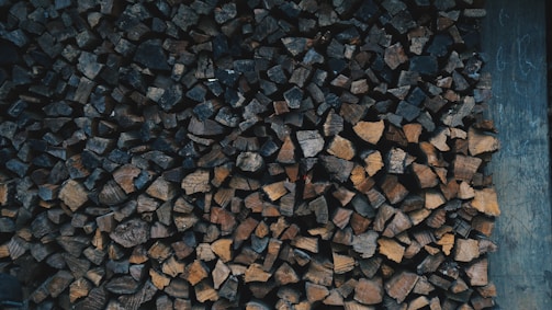 A stacked pile of chopped firewood in various shades of brown, neatly arranged against a wooden backdrop. The firewood pieces are of different shapes and sizes, creating a textured pattern.