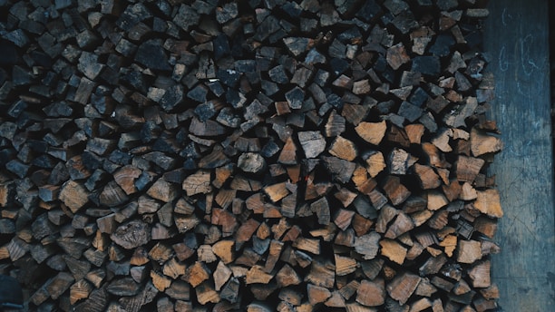A stacked pile of chopped firewood in various shades of brown, neatly arranged against a wooden backdrop. The firewood pieces are of different shapes and sizes, creating a textured pattern.