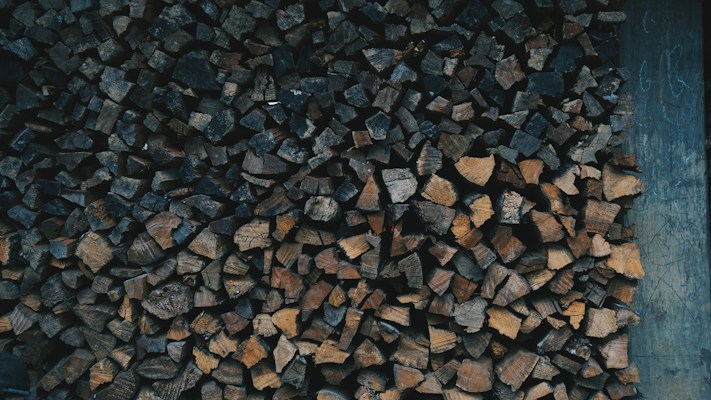 A stacked pile of chopped firewood in various shades of brown, neatly arranged against a wooden backdrop. The firewood pieces are of different shapes and sizes, creating a textured pattern.