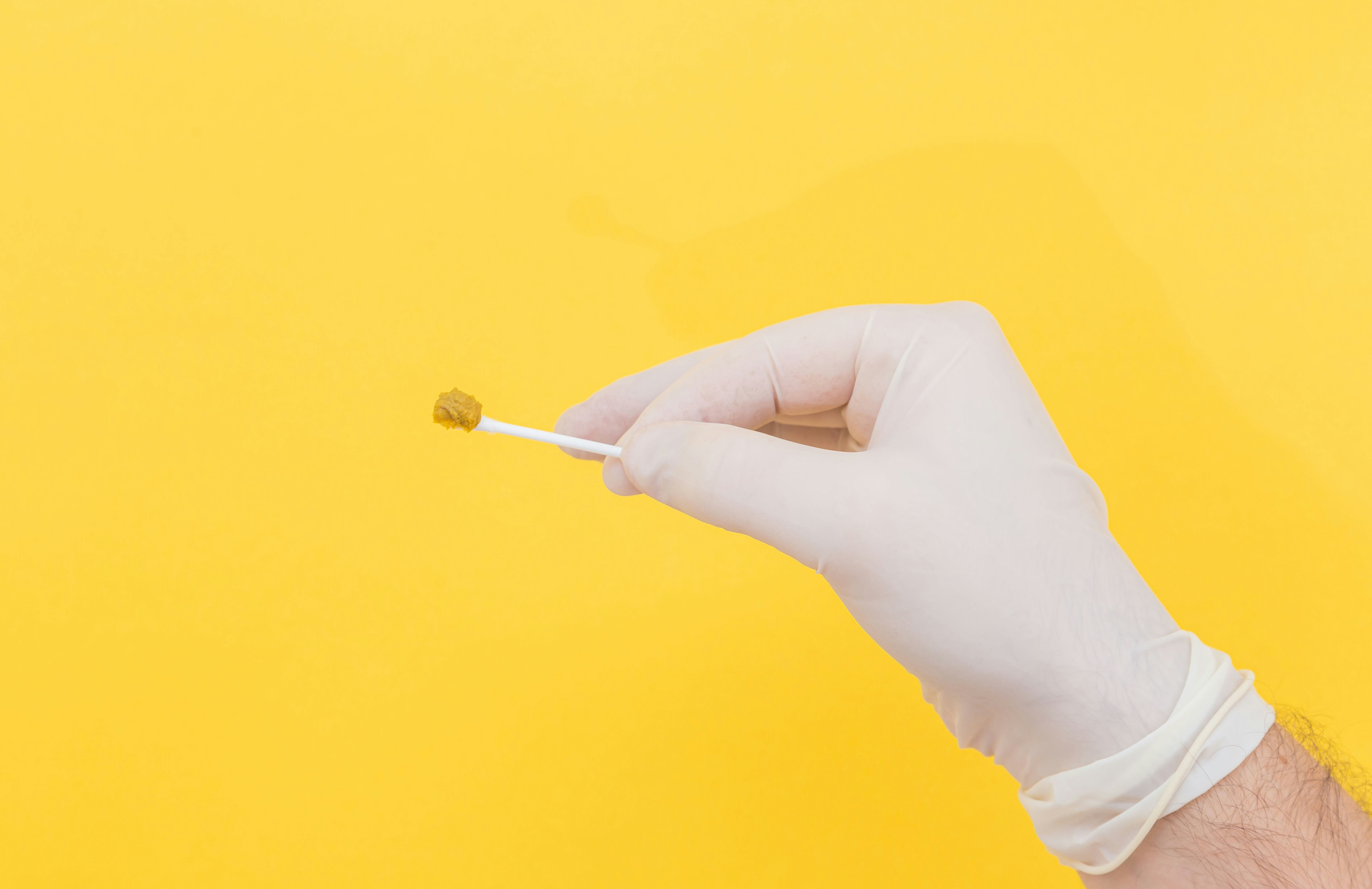 A doctor's hand in a white medical glove holds a stool sample on a stick on a yellow background. The medicine. Analysis. Cala. Infection. The close plan.