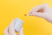 A pair of gloved hands holding a small white container and a swab with a brown substance against a bright yellow background.