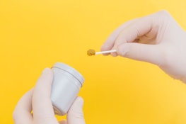 A pair of gloved hands holding a small white container and a swab with a brown substance against a bright yellow background.