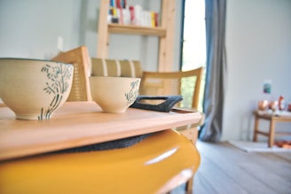 Ceramic cups with a blue floral design are placed on a wooden tray. The background reveals a cozy room with wooden furniture, a shelf with books, and soft lighting from a large window.