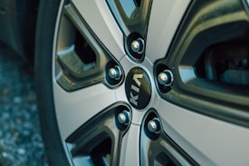 A close-up view of a car wheel with a metallic, multi-spoke design. The center features the KIA logo in white letters on a dark background. The wheel has a shiny, reflective surface with muted metallic tones.