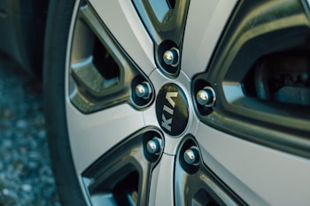 A close-up view of a car wheel with a metallic, multi-spoke design. The center features the KIA logo in white letters on a dark background. The wheel has a shiny, reflective surface with muted metallic tones.