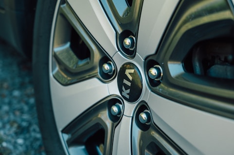 A close-up view of a car wheel with a metallic, multi-spoke design. The center features the KIA logo in white letters on a dark background. The wheel has a shiny, reflective surface with muted metallic tones.