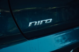 A close-up view of a teal-colored car featuring the silver emblem with the text 'niro' prominently displayed on the rear section. The surrounding surface of the car reflects ambient light, emphasizing its glossy finish.