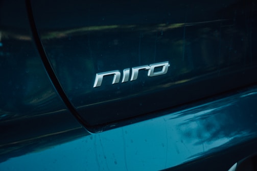 A close-up view of a teal-colored car featuring the silver emblem with the text 'niro' prominently displayed on the rear section. The surrounding surface of the car reflects ambient light, emphasizing its glossy finish.