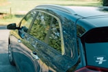 The close-up side view of a car reflects trees and foliage in its windows and glossy surface. The car, parked on a street, displays a sticker indicating a 7-year warranty on the rear window. The background includes a blurred landscape with greenery.