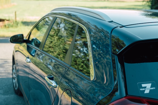 The close-up side view of a car reflects trees and foliage in its windows and glossy surface. The car, parked on a street, displays a sticker indicating a 7-year warranty on the rear window. The background includes a blurred landscape with greenery.