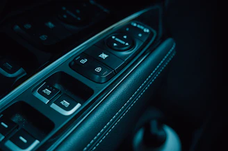 The image displays the controls on a car door panel, including buttons for window adjustments and locking mechanisms. The panel features a sleek design with an emphasis on functionality, highlighted by a stitched leather armrest.