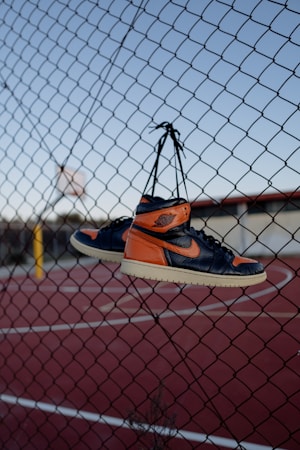 A pair of orange and black sneakers are hanging by their laces from a chain-link fence. The background features a red outdoor basketball court and a clear blue sky.