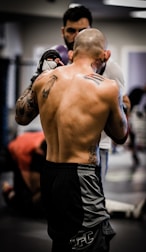 A shirtless, tattooed man with a bald head is engaged in a training or sparring session in an indoor gym environment. He is wearing black and gray sports shorts, and there is a focus on his upper body muscles and tattoos. In the background, another person is visible, possibly a coach or fellow participant, which adds to the dynamic energy of the scene.