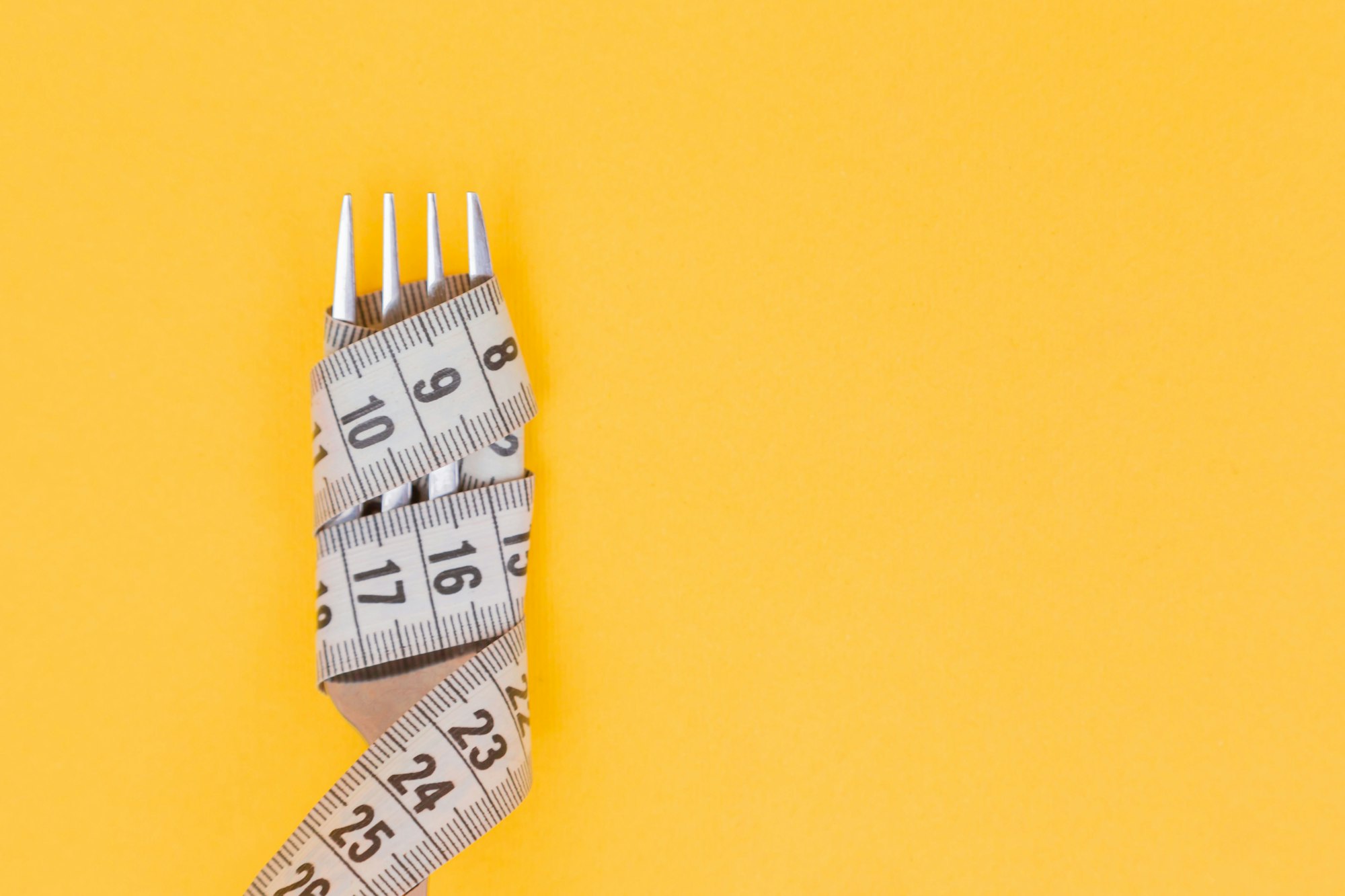 Iron fork and white measuring tape on a yellow background. Diet. Slimming. Obesity. Place for an inscription. Weight loss marathon. Advertising. Top view. The close plan.