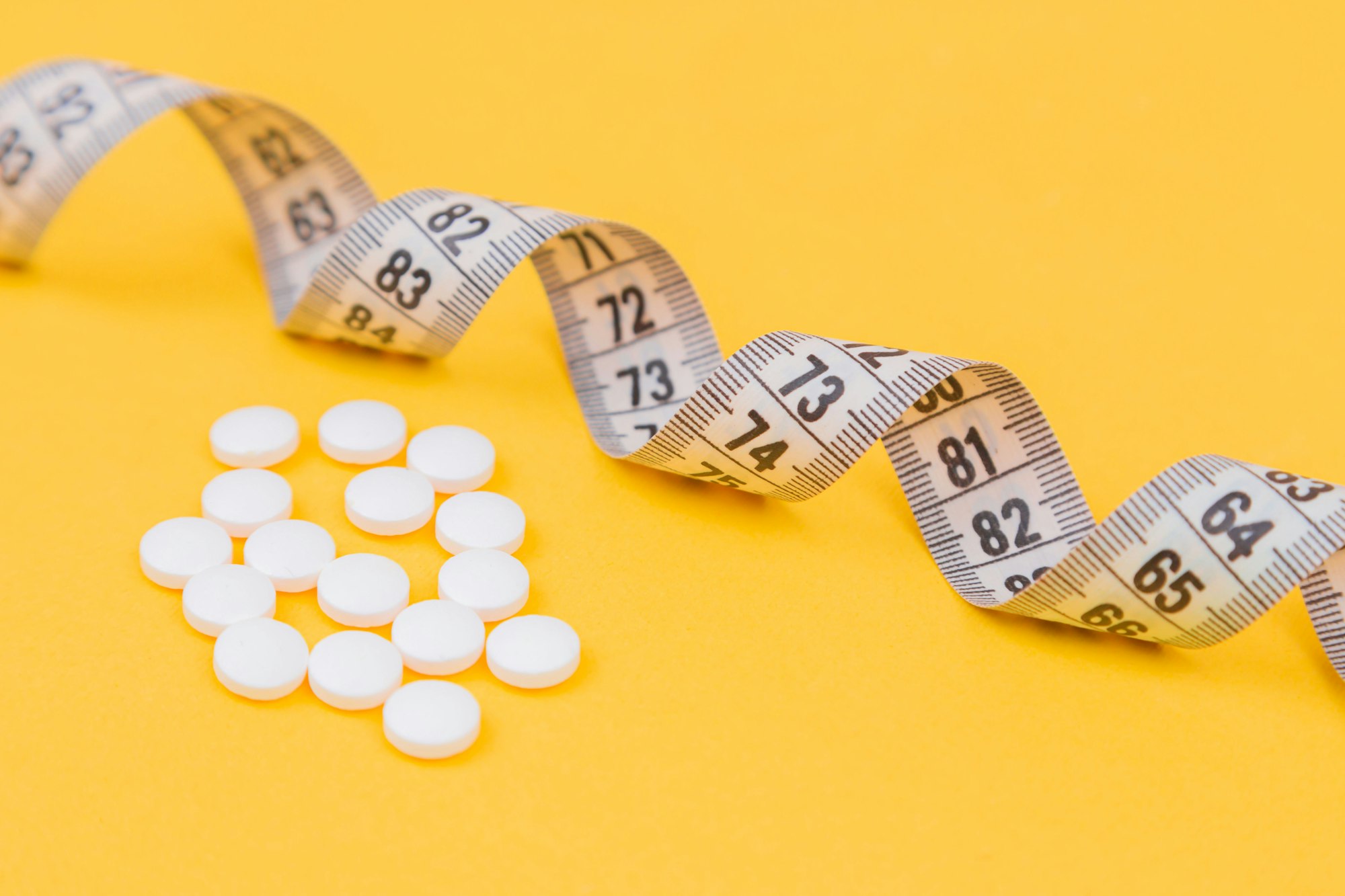 White measuring tape with a white pills, white vitamin on a yellow background. Diet. Slimming. Obesity. Place for an inscription. Weight loss marathon. Advertising. Top view. The close plan.