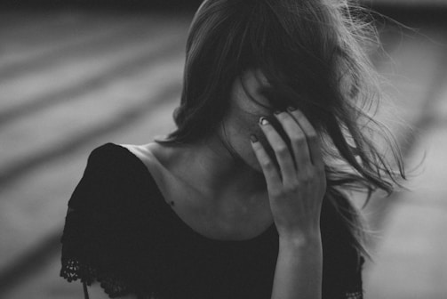 A black and white image of a person with long hair covering their face, wearing a dark top with lace sleeves. The person’s hand is raised to their face, partially obscuring their features. The background consists of blurred steps, suggesting an outdoor setting.