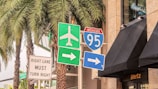 A collection of road signs located at a busy intersection, with an Interstate 95 sign, an airport directional sign, and other traffic instructions. Palm trees and a building with a Hertz sign are in the background.
