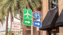 A collection of road signs located at a busy intersection, with an Interstate 95 sign, an airport directional sign, and other traffic instructions. Palm trees and a building with a Hertz sign are in the background.