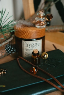 A cozy arrangement featuring a brown glass jar labeled 'hygge' and topped with a wooden lid. A small amber bottle with a cork is nearby, accompanied by a golden bell, a feather, and a green pine branch. A navy blue leather notebook with a brown string and a decorative compass design rests at the bottom. The scene conveys a sense of warmth and comfort.