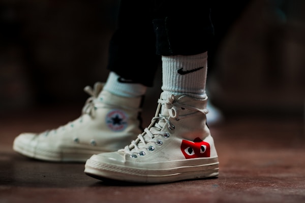 A pair of off-white high-top sneakers with a distinctive red and black design on the side, worn with white socks featuring a black logo. The shoes are on a brown surface, and the lighting creates a moody and stylish atmosphere.