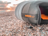 A close-up view of a black running shoe lying on a textured surface, possibly gravel or pebbles. The shoe has the word 'RUNNING' in orange letters along with a small logo. In the background, there is a soft focus of a sunset or sunrise with light clouds.