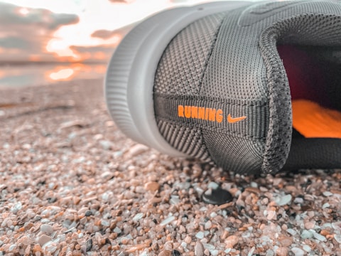 A close-up view of a black running shoe lying on a textured surface, possibly gravel or pebbles. The shoe has the word 'RUNNING' in orange letters along with a small logo. In the background, there is a soft focus of a sunset or sunrise with light clouds.