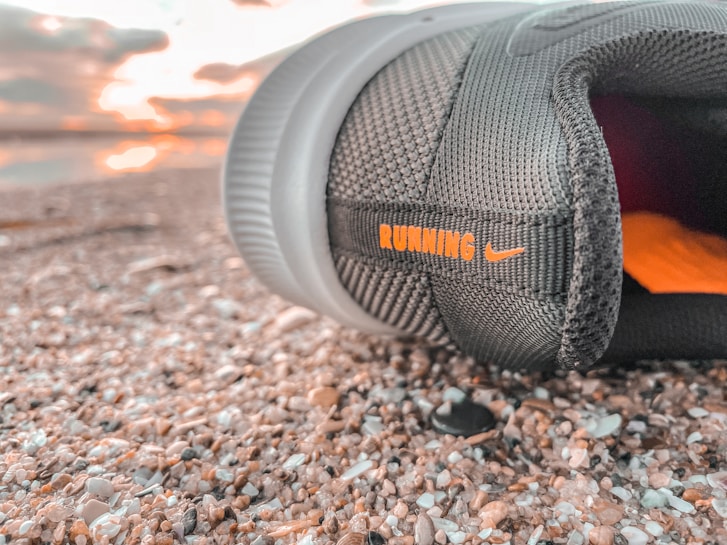 A close-up view of a black running shoe lying on a textured surface, possibly gravel or pebbles. The shoe has the word 'RUNNING' in orange letters along with a small logo. In the background, there is a soft focus of a sunset or sunrise with light clouds.