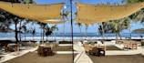 An outdoor dining area on a beachfront with several wooden tables and chairs set under large beige canopies. String lights hang from the canopies, and palm trees are scattered in the background. The ocean is visible beyond the dining area, with waves crashing on the shore and boats in the distance.