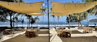 An outdoor dining area on a beachfront with several wooden tables and chairs set under large beige canopies. String lights hang from the canopies, and palm trees are scattered in the background. The ocean is visible beyond the dining area, with waves crashing on the shore and boats in the distance.