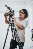 A person with curly hair and glasses is adjusting a professional video camera on a tripod in a bright room. They are focused on the camera, wearing a plain white T-shirt and dark pants. A fan is partially visible in the background.