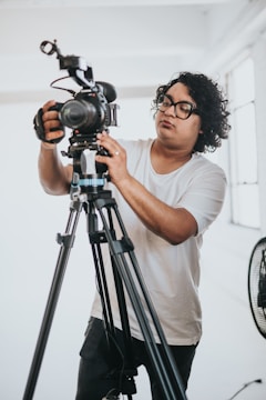 A person with curly hair and glasses is adjusting a professional video camera on a tripod in a bright room. They are focused on the camera, wearing a plain white T-shirt and dark pants. A fan is partially visible in the background.