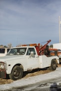 A vintage tow truck with a white cab and a rusty red towing mechanism is parked on a patch of grass and snow. The truck appears weathered, with visible rust and worn paint. In the background, other vehicles and a utility pole can be seen under a clear blue sky.