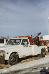 A vintage tow truck with a white cab and a rusty red towing mechanism is parked on a patch of grass and snow. The truck appears weathered, with visible rust and worn paint. In the background, other vehicles and a utility pole can be seen under a clear blue sky.