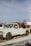 A vintage tow truck with a white cab and a rusty red towing mechanism is parked on a patch of grass and snow. The truck appears weathered, with visible rust and worn paint. In the background, other vehicles and a utility pole can be seen under a clear blue sky.